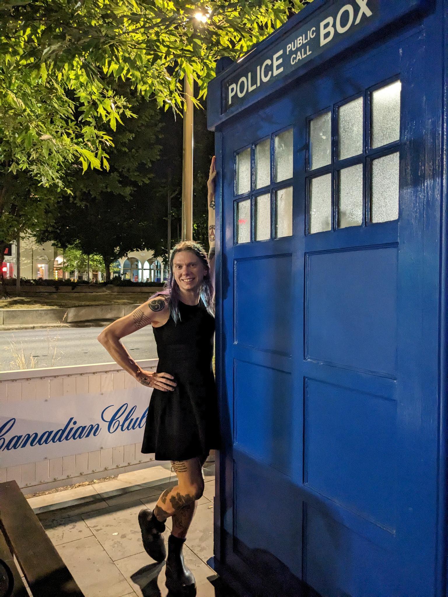 Stacey stands leaning against the TARDIS with one hand  and with one leg behind the other. It's a well-lit tree-lined street. Behind her on a low fence, in a cursive typeface, are the words Canadian Club.