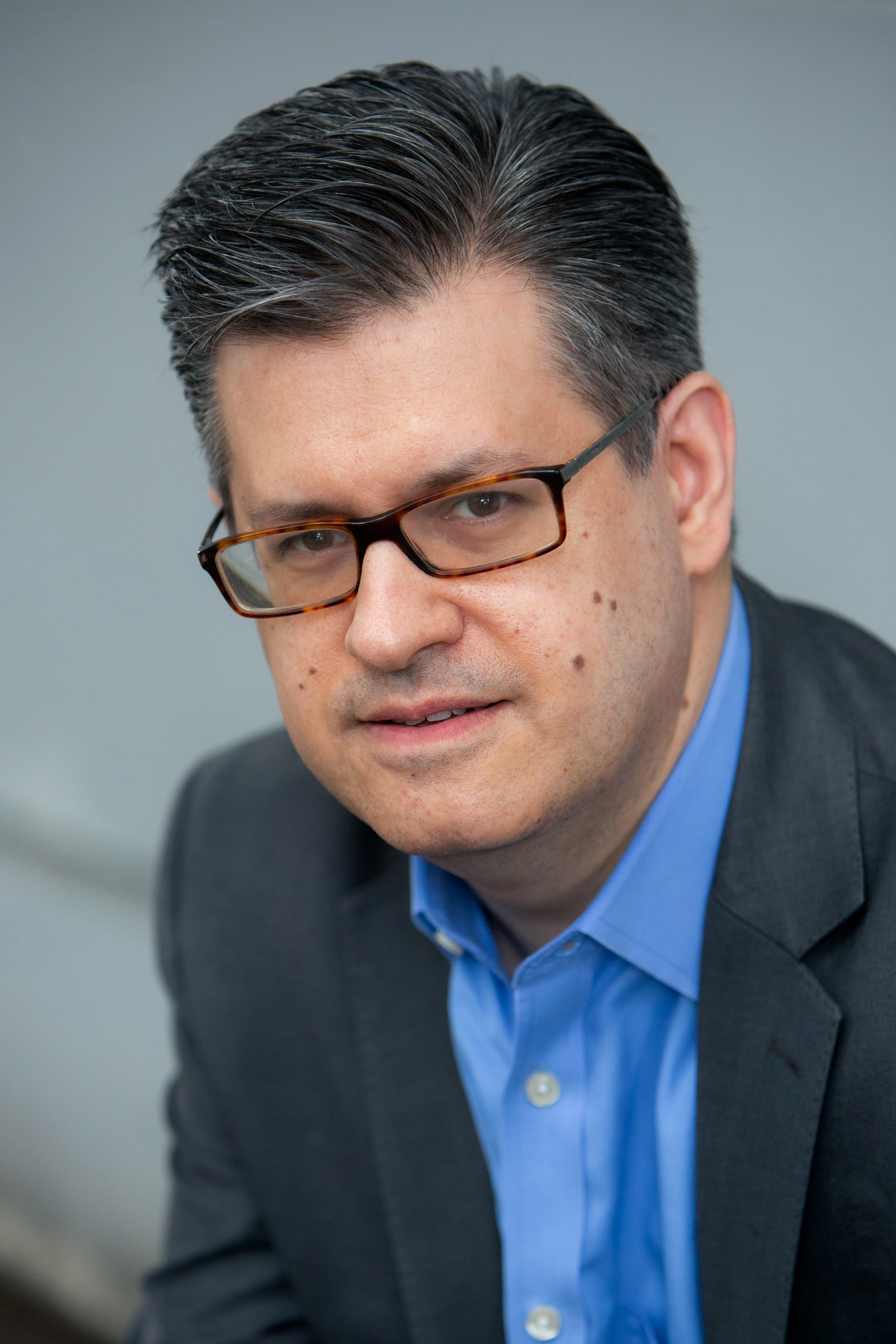 Handsome publicity shot of Tom, wearing glasses, a blue shirt and a grey suit jacket.