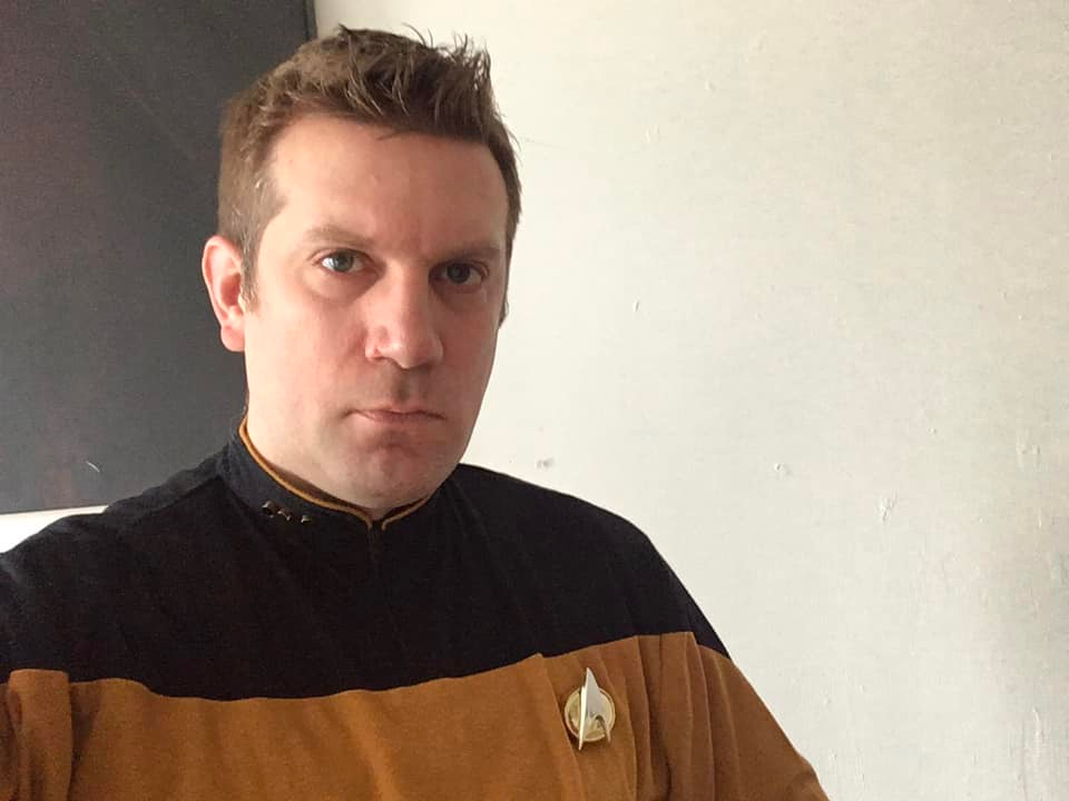 Tom Spilsbury, looking serious, wearing a gold Star Trek The Next Generation uniform.