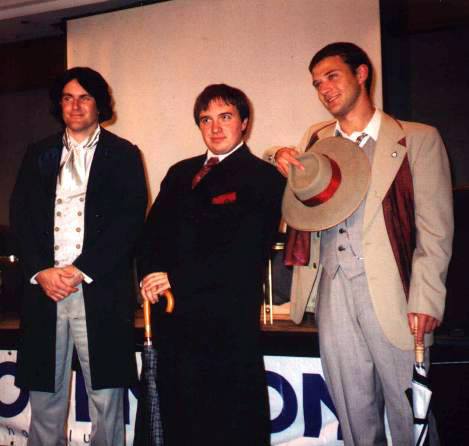 Three young men on stage, one dressed as the Eighth Doctor, one in a dark suit and paisley tie, and one dressed as the Seventh Doctor from Season 24