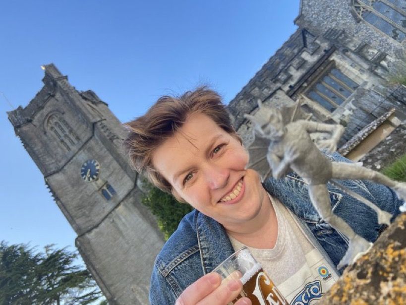 A young woman is drinking a pint out the front of the of the church at Devil's End, with a Bok figure standing on the table in front of her.