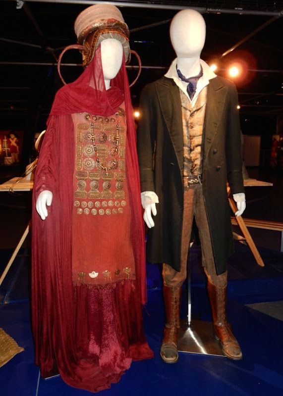 Two mannequins wearing Doctor Who costumes—Ohila’s ornate red robe and headdress, and the Eighth Doctor’s worn 19th-century-style outfit with coat, waistcoat, and boots—on display under exhibit lighting.